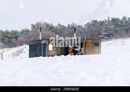 Sauna all'aperto in legno con fumo che esce dal camino in una bella giornata invernale fredda e innevata al Mar Baltico. Benessere e stile di vita sano. Foto Stock