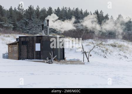 Sauna all'aperto in legno con fumo che esce dal camino in una bella giornata invernale fredda e innevata al Mar Baltico. Benessere e stile di vita sano. Foto Stock