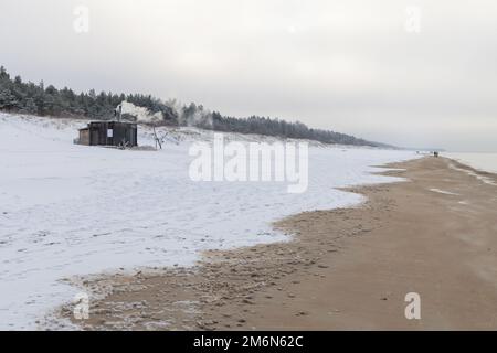 Sauna all'aperto in legno con fumo che esce dal camino in una bella giornata invernale fredda e innevata al Mar Baltico. Benessere e stile di vita sano. Foto Stock