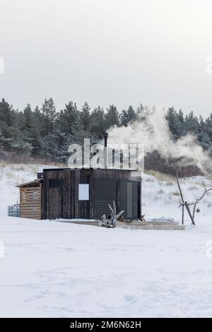 Sauna all'aperto in legno con fumo che esce dal camino in una bella giornata invernale fredda e innevata al Mar Baltico. Benessere e stile di vita sano. Foto Stock