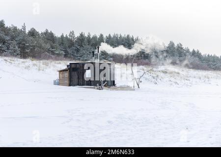 Sauna all'aperto in legno con fumo che esce dal camino in una bella giornata invernale fredda e innevata al Mar Baltico. Benessere e stile di vita sano. Foto Stock