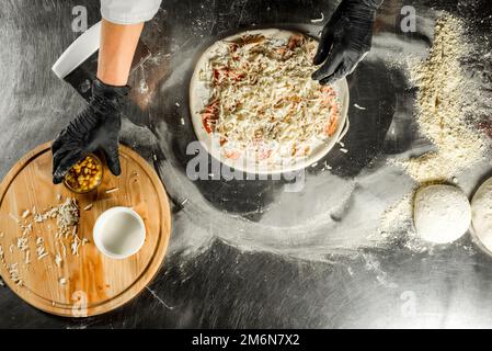 Cucinare la pizza su un tavolo d'acciaio; pasta, farina, farcitura, salsa e le mani dello chef in guanti neri sul tavolo Foto Stock