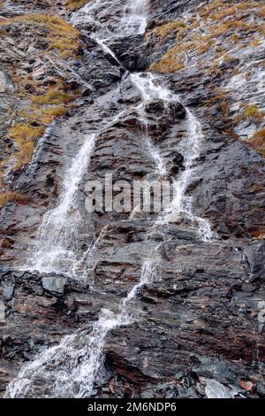 Piccola cascata di montagna scorre su creste granitiche di rocce alpine ricoperte di muschi e licheni autunnali ingialliti, tiro verticale, Cogne, Valle d'Aosta Foto Stock
