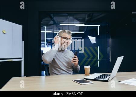 Capo d'affari dai capelli grigi senior che lavora all'interno di un ufficio moderno utilizzando un computer portatile al lavoro, uomo maturo in cuffie che ascolta musica e audiolibri podcast utilizzando l'app sul telefono. Foto Stock