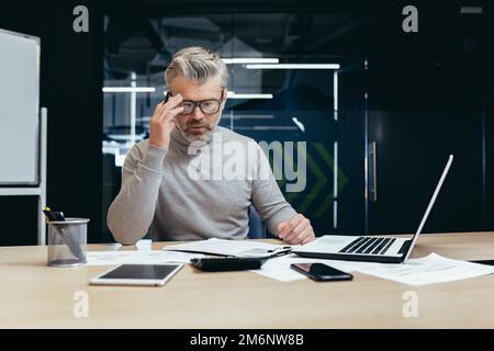 Uomo d'affari senior che lavora in ufficio con notebook, uomo che pensa a decisioni finanziarie importanti. Foto Stock