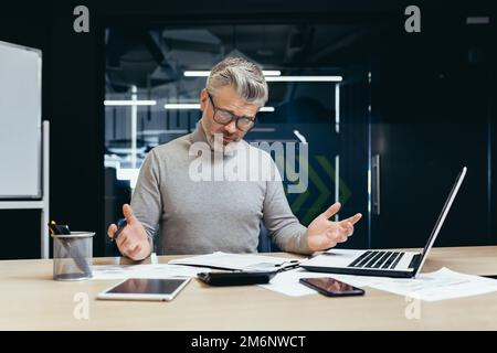 Boss dai capelli grigi insoddisfatto in ufficio, uomo anziano deluso dal risultato ottenuto, uomo d'affari maturo turbato e triste lavorare con i documenti e utilizzare il laptop all'interno dell'edificio. Foto Stock