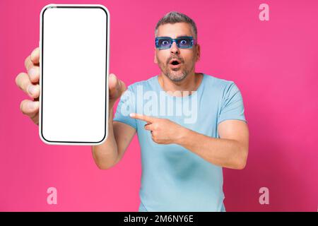 Scioccato bell'uomo 40s che indica il grande smartphone in mano indossando una camicia blu e occhiali da sole isolati su sfondo rosa. Uomo w Foto Stock