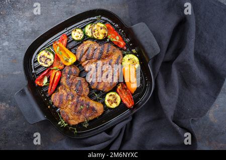 Tradizionale bistecca di maiale alla griglia con verdure alla griglia servita e spezie in primo piano in una padella dal design rustico con cop Foto Stock