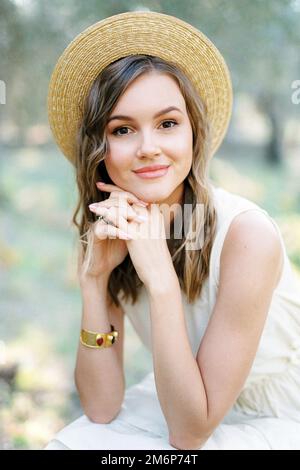 Una giovane donna in un cappello di paglia si siede con le mani attaccate sotto il mento Foto Stock