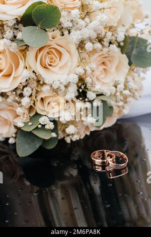 Una bella immagine dai toni con gli anelli di nozze si trova su una superficie nera contro lo sfondo di un bouquet di fiori Foto Stock