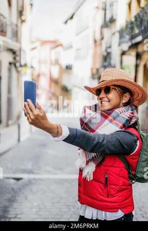 una donna di mezza età che indossa un cappello e occhiali da sole, prende un selfie per strada mentre sorride. concetto di turismo e felicità. Foto Stock