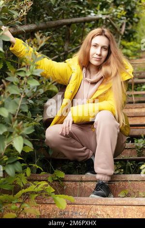 Una donna bionda in una giacca gialla è seduta sulle scale. Scale vecchie troppo grandi Foto Stock