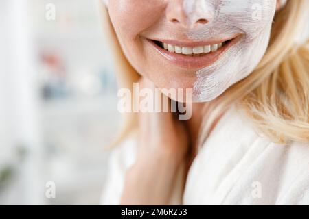 Primo piano scatto di donna irriconoscibile con maschera bianca crema su una metà del suo volto è sorridente Foto Stock
