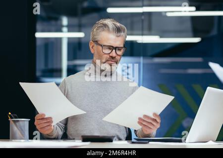 Frustrato e arrabbiato uomo d'affari con i documenti, maturo grigio uomo d'affari che controlla i rapporti finanziari, capo non soddisfatto con i risultati di realizzazione e reddito che lavora all'interno dell'ufficio con il laptop. Foto Stock