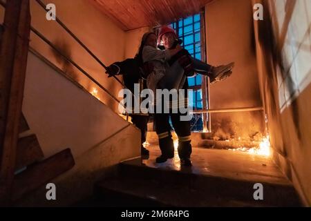 Il Fireman coraggioso scende le scale di un edificio che brucia e tiene la ragazza salvata nelle sue braccia. Fuoco aperto e un pompiere nel backg Foto Stock