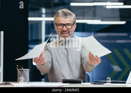 Frustrato e arrabbiato uomo d'affari con i documenti, maturo grigio uomo d'affari che controlla i rapporti finanziari, capo non soddisfatto con i risultati di realizzazione e reddito che lavora all'interno dell'ufficio con il laptop. Foto Stock