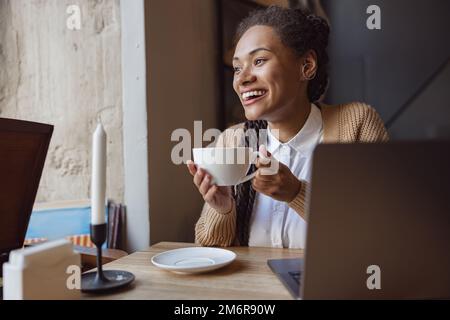 Sorridente bella giovane donna africana, studente, blogger con computer portatile e cappuccino tazza da solo in un accogliente bar Foto Stock