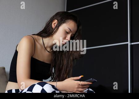 Ragazza teen con lunghi capelli seduti in pigiama con smartphone a letto. Comunicazione online, tempo libero a casa Foto Stock