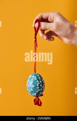 Mano che tiene un uovo di Pasqua blu decorato con cera di fronte a sfondo arancione in primavera Foto Stock