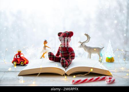 Libro di carta aperto con bella figura orsacchiotto forma orso, luci fata illuminate su tavolo bianco, paesaggio boschivo nevoso sullo sfondo. Foto Stock