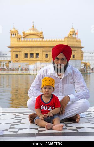 Sikh indiano in vestito bianco e a piedi nudi con il figlio seduto al bordo del bacino d'acqua dell'Amrit Sagar o Lago Sacro, presso l'Hari Mandir OR Foto Stock