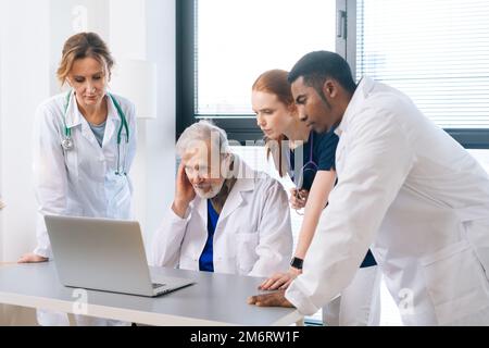 Un team medico serio di medici multietnici che parlano insieme concentrato guardando allo schermo del computer portatile in piedi sullo sfondo della finestra. Foto Stock