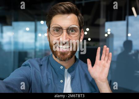 Ritratto di un uomo d'affari di successo in una camicia casual e occhiali uomo che guarda la fotocamera dello smartphone e sorridente, vista fotocamera videochiamata parlare con gli amici in ufficio in remoto. Foto Stock