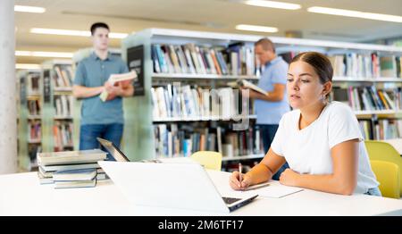 Giovane donna concentrata che controlla la pianificazione durante lo studio e l'uso del computer portatile in biblioteca Foto Stock