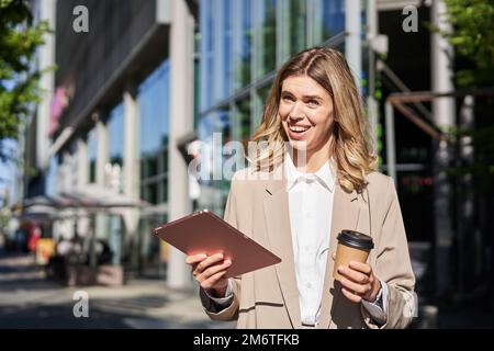 Ritratto di giovane donna d'affari fiduciosa sulla strada, beve il suo caffè e guarda il tablet, lavora sulla sua strada per l'ufficio. Digitale Foto Stock