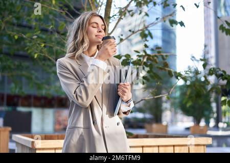 Donna sorridente in tuta, bevendo caffè da asporto sulla strada vicino all'edificio degli uffici Foto Stock