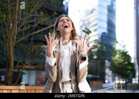 Entusiasta donna d'affari gioire, celebrare e sentirsi felice, esprimere pura gioia, in piedi per strada in vestito beige Foto Stock