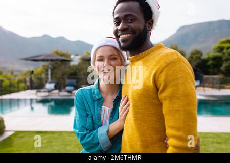 Ritratto di felice coppia diversa in cappelli di santa per celebrare il natale sorridendo all'aperto Foto Stock