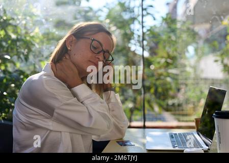 Donna stressata stanca che soffre di dolore alla nuca lavorando da casa ufficio seduto a tavolo. Donna giovane anziana iperlavorata massaggiando il collo sensazione di dolore ferito Foto Stock