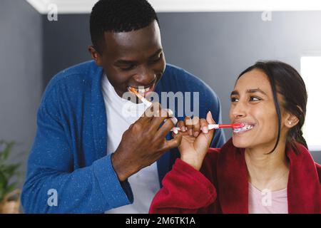 Felice coppia diversa nel spazzolare i denti in bagno, guardandosi sorridendo Foto Stock