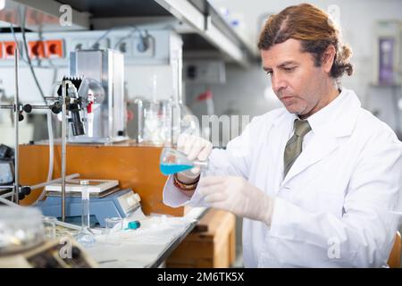 Biochimico latino-maschile professionale che studia il comportamento dei reagenti liquidi blu mentre lavora in un laboratorio moderno Foto Stock