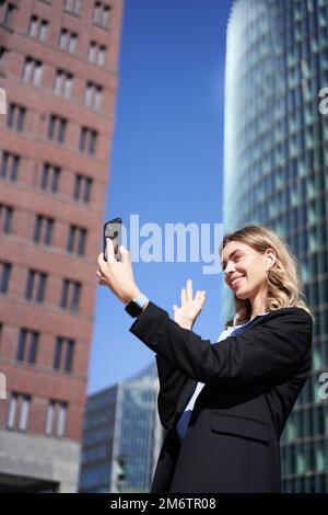 Ritratto della sorridente videochiamata aziendale di una donna in strada, tenendo in mano il telefono cellulare e muovendo la fotocamera dello smartphone, indossando la tuta Foto Stock
