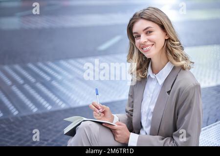 Donna in tuta si siede nel centro della città, scrive, prende appunti, tiene la penna e il notebook, brainstorms, crea idee Foto Stock