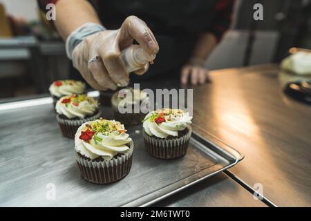 Processo di decorazione dei cupcake. Chiudi il colpo di molti cupcake dolci in primo piano mentre un panettiere decorare uno di loro con gli spruzzi. Foto di alta qualità Foto Stock