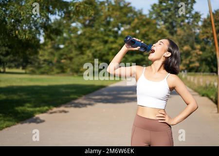 Buon sportivo asiatico, runner beve acqua dalla bottiglia mentre correndo, allenarsi all'aria fresca nel parco Foto Stock