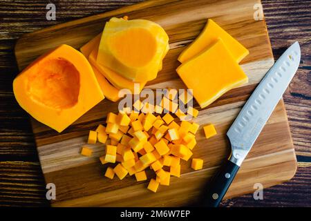 Zucca di noce taglio in piccoli cubetti su un tagliere di legno: Zucca di inverno a dadini su uno sfondo di legno con un coltello da cucina Foto Stock