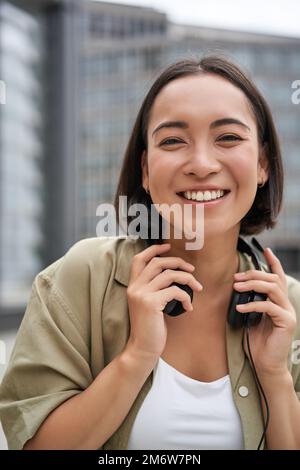 Foto verticale di una bella donna asiatica che si posa con le cuffie intorno al collo, sorridente e ridente, in piedi per strada alla luce del giorno Foto Stock