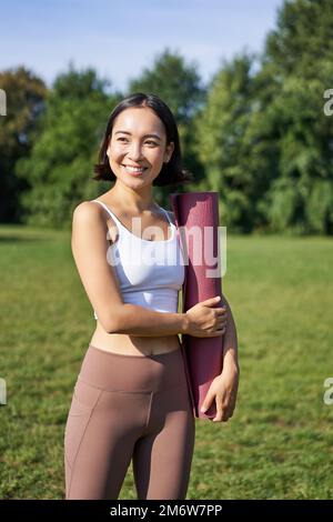 Ritratto verticale di giovane ragazza asiatica fitness passeggiate con tappetino di gomma per lo yoga, va in sessione di allenamento all'aria fresca nel parco, Wea Foto Stock