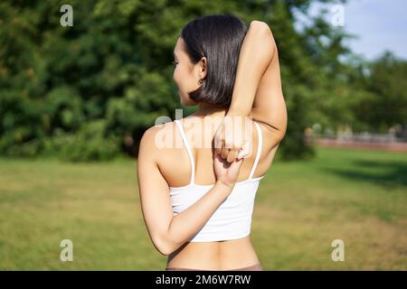 Vista posteriore della giovane donna sportiva che si stende le braccia dietro la schiena, riscaldamento, preparazione per il jogging allenamento, evento sportivo nel parco Foto Stock