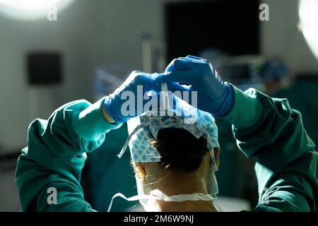 Vista posteriore di chirurgo in cappuccio chirurgico e guanti che legano la maschera in sala operatoria Foto Stock