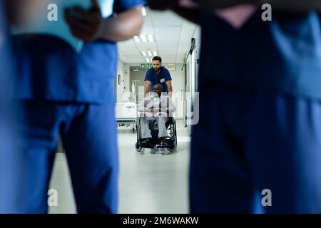 Diversi operatori sanitari maschili che spingono il paziente maschio afro-americano in sedia a rotelle in ospedale Foto Stock