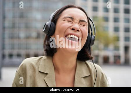 Ritratto di felice donna asiatica in cuffie, ama la musica mentre cammina in città, sorridendo e ridendo Foto Stock