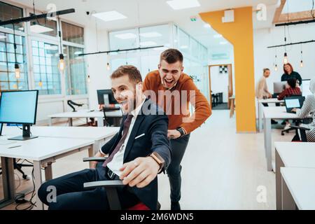 Team building e divertimento in ufficio. Giovane uomo d'affari allegro in abbigliamento casual elegante che si diverte mentre gareggiare su sedie da ufficio e SM Foto Stock