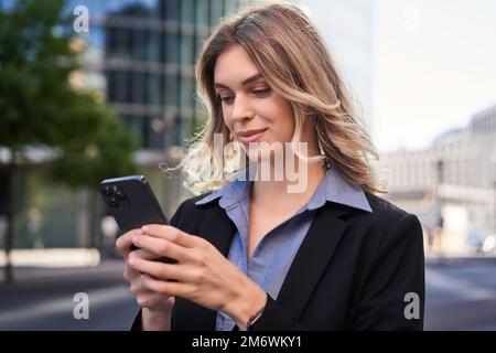 Primo piano ritratto di una giovane donna aziendale in tuta nera, tiene in mano smartphone, SMS mentre si è in piedi per strada, utilizzando mobi Foto Stock