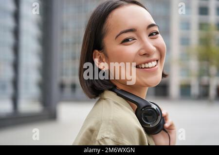 Ritratto di felice donna asiatica in cuffie, ama la musica mentre cammina in città, sorridendo e ridendo Foto Stock