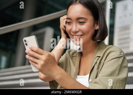 Ritratto di ragazza asiatica prende selfie sul telefono cellulare. Donna coreana sorridente, videochattare sull'app per smartphone mentre si siede all'esterno di st Foto Stock